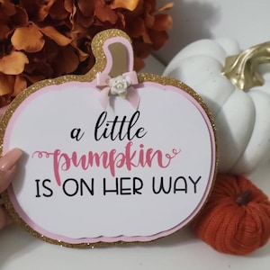 Może przedstawiać: Tablica w kształcie dyni z napisem "a little pumpkin is on her way". Tablica ma r&oacute;żowo-złotą brokatową obw&oacute;dkę i małą kokardkę. Biała dynia i pomarańczowe kwiaty są w tle.
