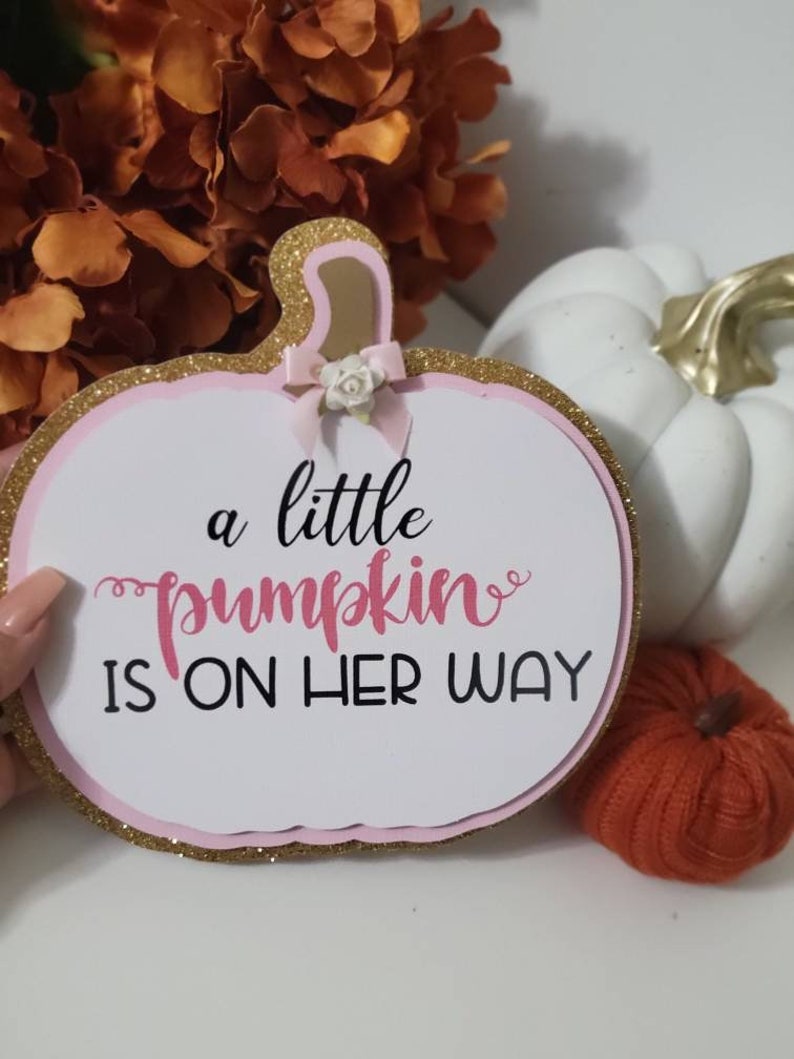 Może przedstawiać: Tablica w kształcie dyni z napisem "a little pumpkin is on her way". Tablica ma r&oacute;żowo-złotą brokatową obw&oacute;dkę i małą kokardkę. Biała dynia i pomarańczowe kwiaty są w tle.