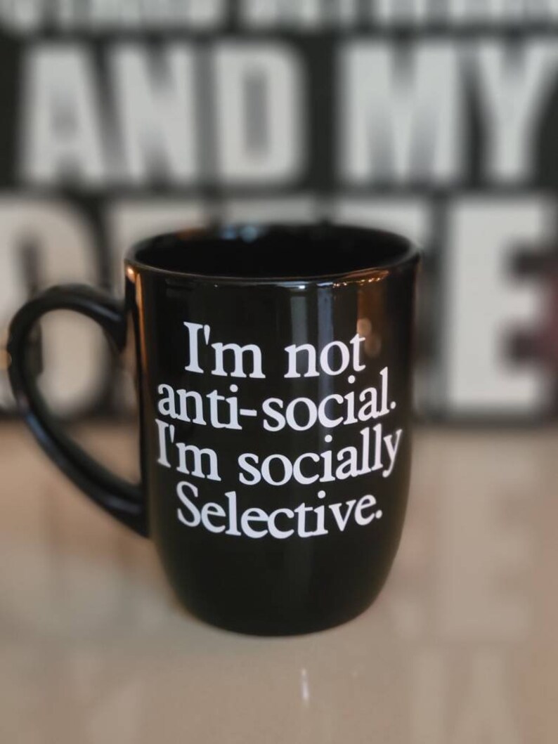 Op de afbeelding: Zwarte koffiemok met witte tekst die luidt: "Ik ben niet anti-sociaal. Ik ben sociaal selectief."