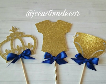 May include: Three gold glitter cake toppers with blue bows. The toppers are shaped like a crown, a onesie, and a diaper. The toppers are perfect for a baby shower or birthday party.