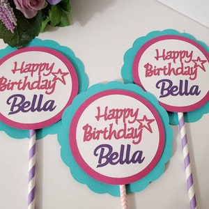 May include: Three turquoise and pink cake toppers with scalloped edges. Each topper has a white circle with the text "Happy Birthday Bella" in pink and purple lettering with a star.