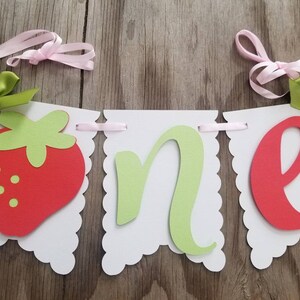 May include: A white banner with scalloped edges, decorated with red and green felt cutouts. The banner spells out "one" with a strawberry on the left side. The banner is strung with pink and green ribbon.