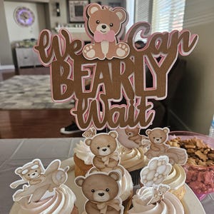 May include: A set of cupcakes decorated with brown teddy bear toppers. The cupcakes are frosted with white frosting and have pink accents. A brown wooden sign with the text "We can bearly wait" is in the background.