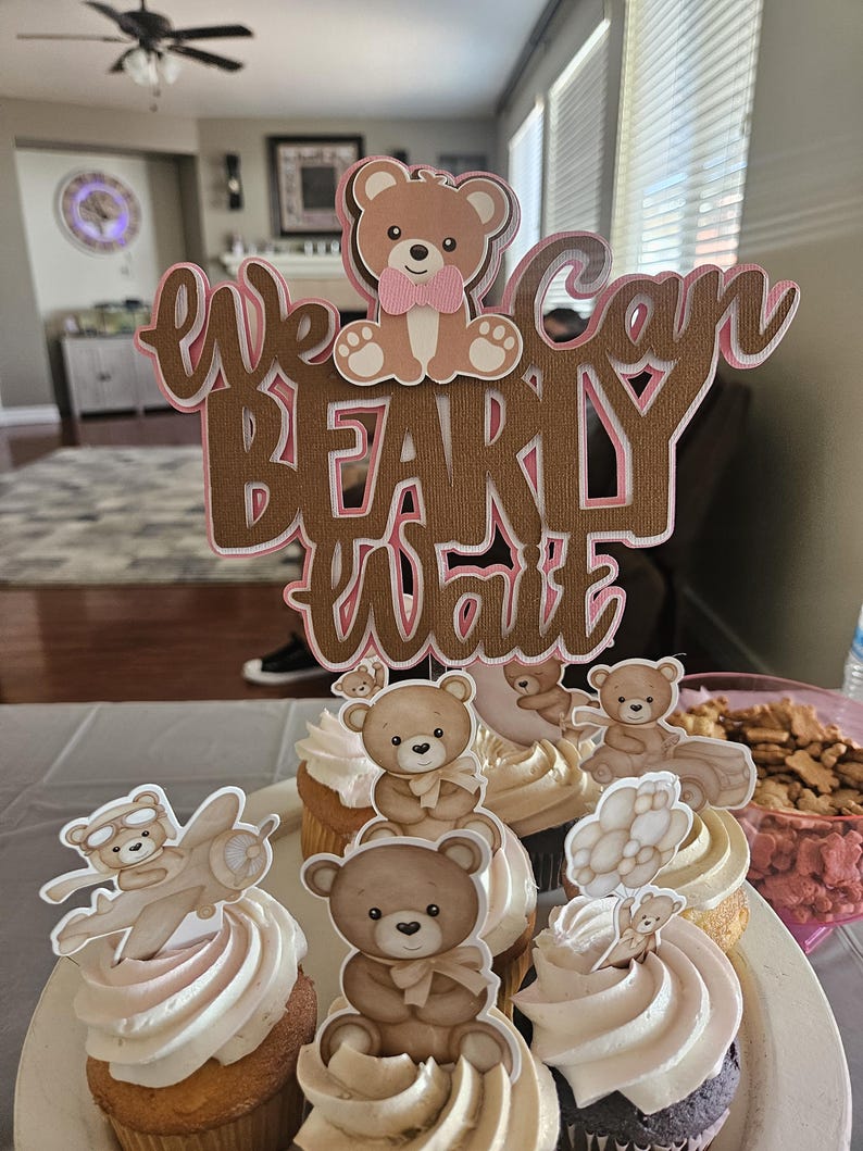 May include: A set of cupcakes decorated with brown teddy bear toppers. The cupcakes are frosted with white frosting and have pink accents. A brown wooden sign with the text "We can bearly wait" is in the background.