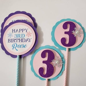 May include: A collection of birthday cake toppers. The toppers feature a purple scalloped edge, with a pink and white circle in the center. One topper says "Happy 3rd Birthday Reese" with snowflake accents. The other toppers have a purple glittery number 3.