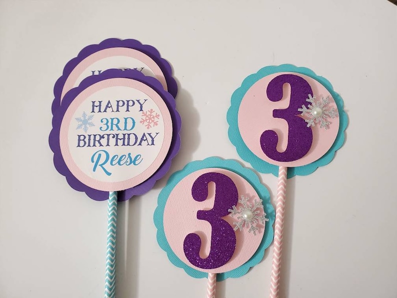 May include: A collection of birthday cake toppers. The toppers feature a purple scalloped edge, with a pink and white circle in the center. One topper says "Happy 3rd Birthday Reese" with snowflake accents. The other toppers have a purple glittery number 3.