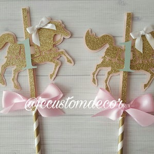 May include: Two gold glitter carousel horse cake toppers with pink bows and a green number one on each. The toppers are on gold and white striped sticks.
