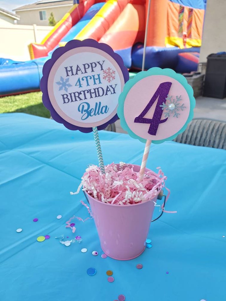 May include: A pink metal bucket filled with pink and white shredded paper, holding two decorative signs. One sign reads "HAPPY 4TH BIRTHDAY Bella" and the other has the number "4". The signs are on striped straws, perfect for a birthday party.