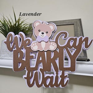 May include: A brown and purple cake topper with the text "We can barely wait" and a cartoon teddy bear wearing a purple bow tie. The topper is on a clear plastic stick.