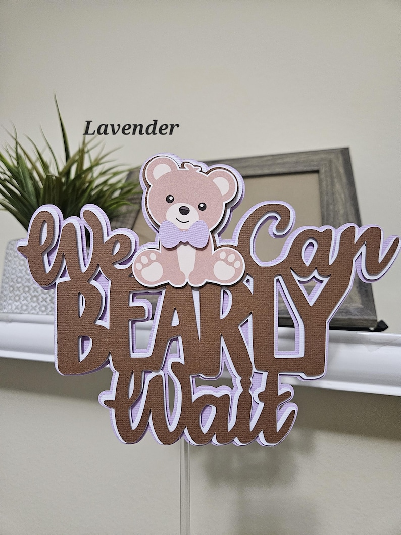 May include: A brown and purple cake topper with the text "We can barely wait" and a cartoon teddy bear wearing a purple bow tie. The topper is on a clear plastic stick.