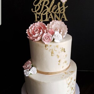 May include: A two-tiered white cake decorated with gold leaf and pink and white sugar flowers. The top tier has a gold glitter topper that says "God Bless Eliana" with a pink cross.