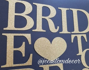 May include: Gold glitter letters spelling "Bride to Be" with a gold glitter heart in the center on a dark blue background. The text "@jccustomdecor" is below the heart.