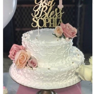 May include: A two-tiered white cake decorated with white icing and a gold glitter cake topper that says "God Bless Sophia" with a pink cross on top. The cake is decorated with pink and peach roses.