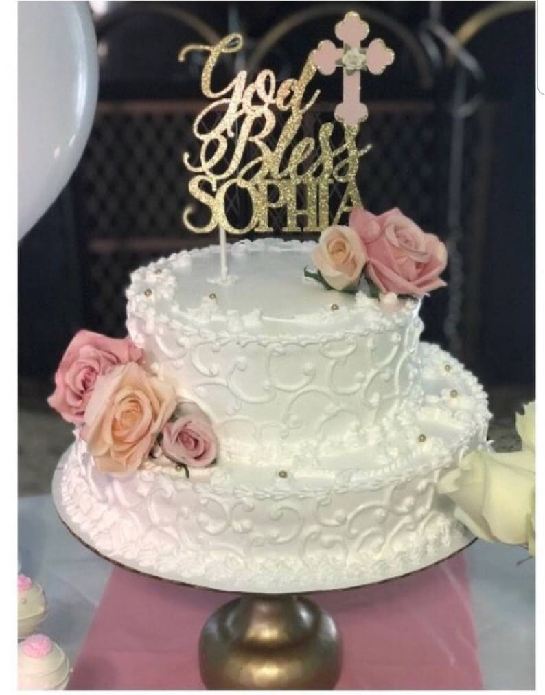 May include: A two-tiered white cake decorated with white icing and a gold glitter cake topper that says "God Bless Sophia" with a pink cross on top. The cake is decorated with pink and peach roses.