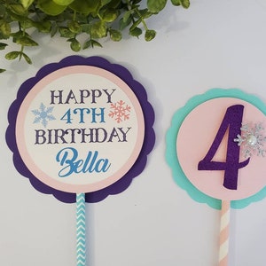 May include: Two birthday cake toppers. One has the text "HAPPY 4TH BIRTHDAY Bella" with snowflakes. The other has a purple number 4 with a snowflake. Both are on sticks with decorative paper.