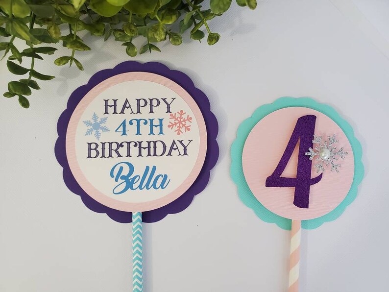May include: Two birthday cake toppers. One has the text "HAPPY 4TH BIRTHDAY Bella" with snowflakes. The other has a purple number 4 with a snowflake. Both are on sticks with decorative paper.
