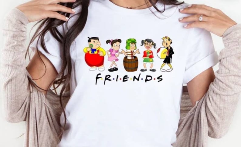 May include: White t-shirt with a cartoon illustration of six friends. The characters are wearing colorful clothing and holding various objects. The text "FRIENDS" is printed below the characters.
