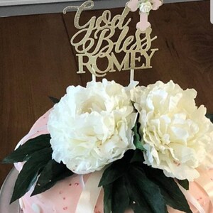 May include: A pink and white cake decorated with white flowers and green leaves. The cake has white polka dots and a gold glitter cake topper that says "God Bless Romey" with a pink cross.