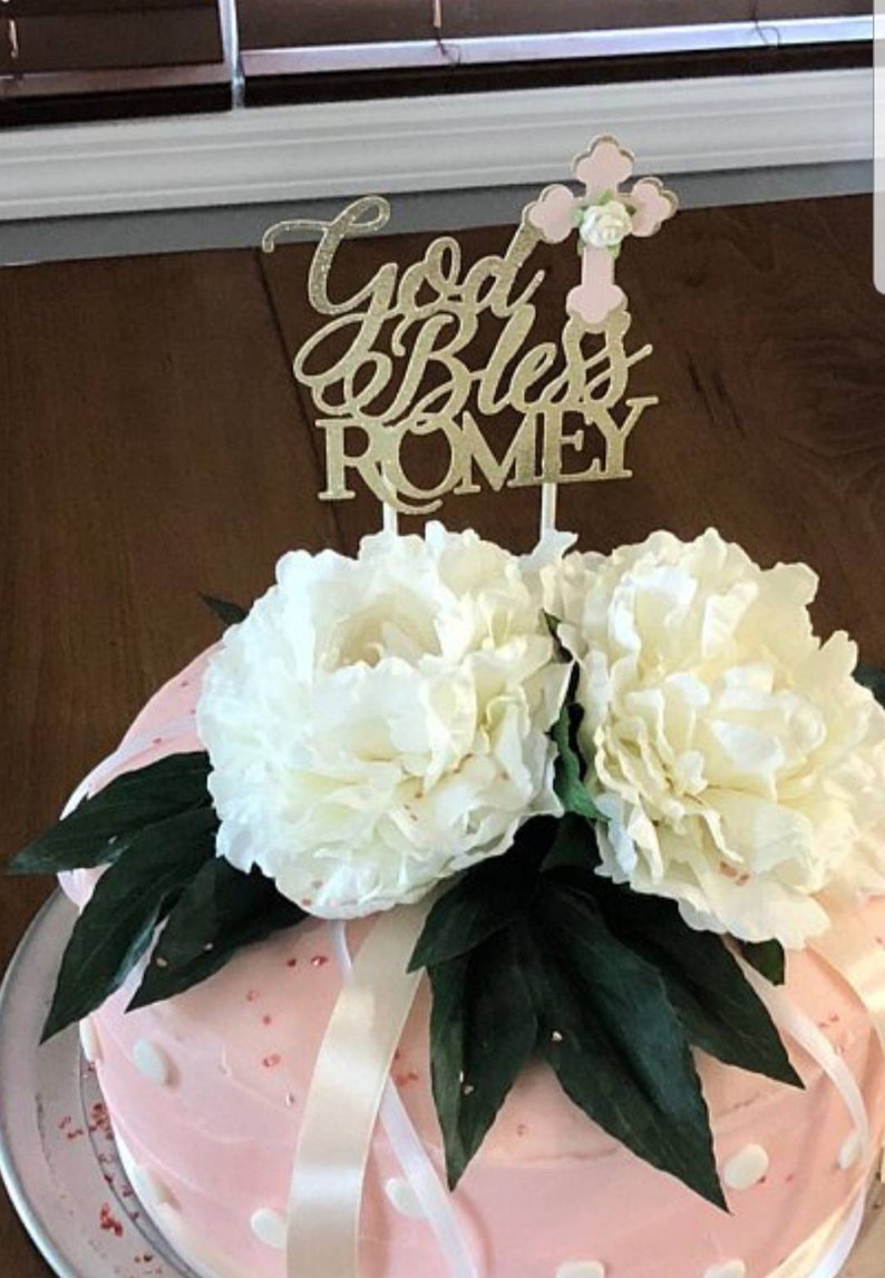 May include: A pink and white cake decorated with white flowers and green leaves. The cake has white polka dots and a gold glitter cake topper that says "God Bless Romey" with a pink cross.