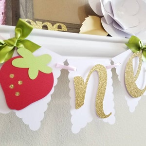 May include: A white and pink banner with the word "one" in gold glitter letters. The banner also features a red and green felt strawberry.