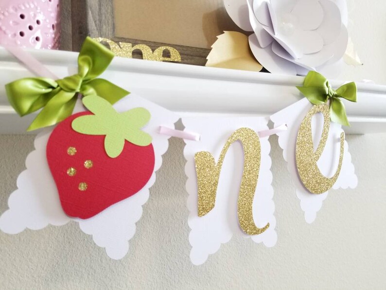 May include: A white and pink banner with the word "one" in gold glitter letters. The banner also features a red and green felt strawberry.