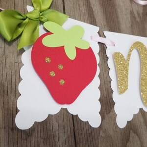 May include: A white banner with a red strawberry, green leaves, and gold glitter letters spelling "one". The banner is decorated with pink and green ribbon.