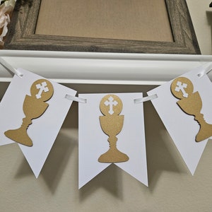 May include: White banner with three gold chalice and cross designs. The banner is strung with white ribbon.