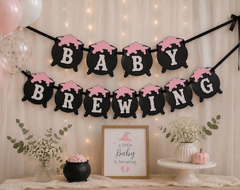 May include: A two-tier banner featuring black cauldron-shaped cutouts with pink bubbling liquid and white letters spelling BABY BREWING, accompanied by a framed sign with the text a little baby is brewing and pink baby footprints.