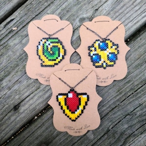 May include: Three pixel art necklaces on brown card stock with the text "Made with Love". The necklaces feature a green and yellow swirl, a blue and yellow design, and a red and yellow heart.