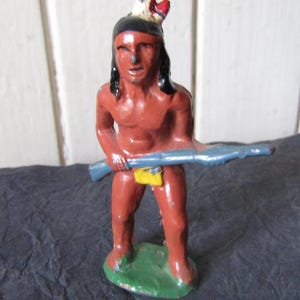 May include: A vintage figurine of a Native American man, painted in shades of red and brown, holding a blue rifle. The figure has black hair and a feathered headdress. It stands on a green base.
