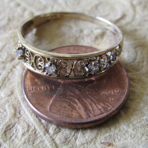 May include: A gold band ring with a delicate filigree design and small clear stones. The ring is placed on top of a US penny for size comparison.