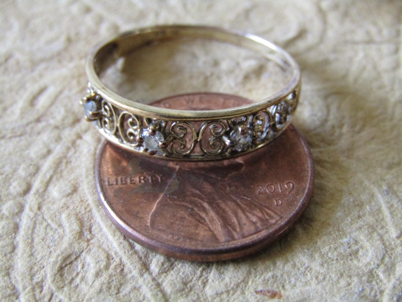 May include: A gold band ring with a delicate filigree design and small clear stones. The ring is placed on top of a US penny for size comparison.