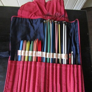 May include: A red fabric roll-up case with a blue lining holds a set of colorful knitting needles. The needles are labeled with numbers 1 through 6.