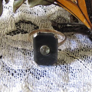May include: A vintage ring with a rectangular black onyx stone and a small, round, clear gemstone in the center. The ring is set on a gold band and rests on a white lace surface. A decorative butterfly is in the background.