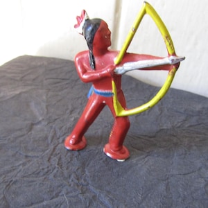 May include: A red toy figure of a Native American archer with a yellow bow and arrow. The figure is painted with black hair, a blue belt, and white accents. The toy is standing on a dark surface.
