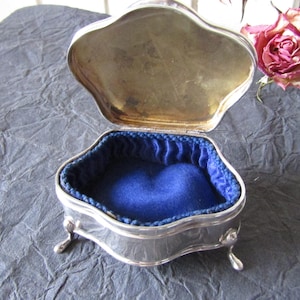 May include: A silver-toned metal trinket box with a hinged, scalloped lid and three small curved legs. The interior is lined with deep blue fabric, featuring a soft, padded base and pleated sides. The metal surface has a polished finish.