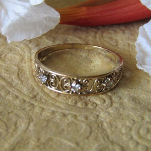 May include: A gold band ring with a delicate filigree design and five small clear stones.