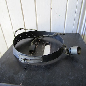 May include: A vintage medical head lamp with a black leather headband and a metal light fixture. The headband has a metal buckle and a series of holes for adjustment. The light fixture has a metal arm and a white porcelain socket.