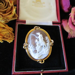 May include: A gold cameo brooch with a white cameo depicting two women in a landscape setting. The cameo is set in a gold frame with a decorative design. The brooch is in a red velvet box with a white label that reads "Hallmarked 9ct Gold, The Carats, 24, Old Bond Street, London, W1, England".