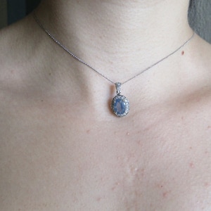 May include: A silver chain necklace with a pendant featuring a large oval-shaped blue gemstone surrounded by smaller clear gemstones.