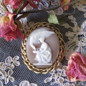 Può includere: Una spilla cammeo ovale con un rilievo bianco scolpito raffigurante una figura alata seduta che interagisce con un cigno su sfondo marrone chiaro, racchiusa in una cornice di metallo dorato con bordo intrecciato.