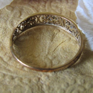 May include: A gold band ring with a delicate, intricate design. The ring is made of a thin band with a filigree pattern.