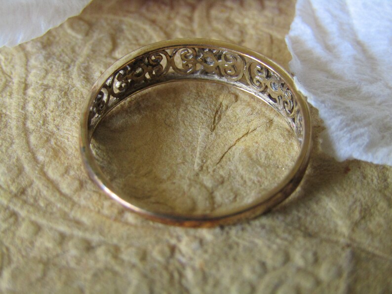 May include: A gold band ring with a delicate, intricate design. The ring is made of a thin band with a filigree pattern.