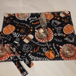 May include: Black placemat and napkins featuring coffee-related designs, such as pumpkins and coffee cups, with text. A silver fork and spoon rest on the placemat. The napkins are folded and placed on the left.