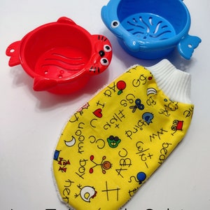 May include: Two colourful plastic bath toys shaped like fish with strainers. The blue fish has a white rim and the red fish has a white rim and black eyes. A yellow fabric washcloth with a white border and black and red drawings of letters, numbers, and other objects.