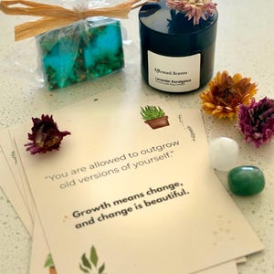 May include: A gift set featuring a teal and green soap bar, a black candle with the text "Affirmed Scents Lavender Eucalyptus", and affirmation cards. The cards have the text "You are allowed to outgrow old versions of yourself." and "Growth means change, and change is beautiful."