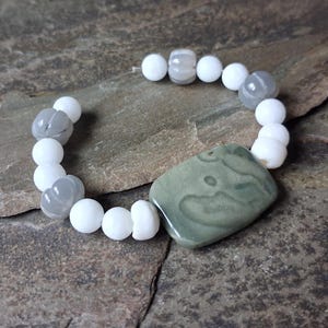 May include: A bracelet featuring a large, rectangular, sage green stone bead with natural swirling patterns, accented by a series of round, matte white beads and translucent, grey, ribbed, pumpkin-shaped beads strung on a clear elastic cord.
