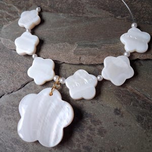 May include: A delicate necklace featuring white, flower-shaped pendants and small pearls. The necklace has a gold-colored clasp and is strung on a clear cord. The flower pendants have a pearlescent sheen.