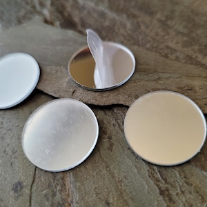 May include: Four small, round, silver-colored, reflective stickers are displayed on a textured, gray-brown surface. One sticker has a protective film partially peeled back, revealing its mirrored surface. The other stickers have a matte finish.