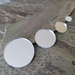 May include: A collection of mirrored, circular craft supplies in varying sizes. The reflective surfaces are silver, and the edges are clean-cut. The items are arranged on a textured, gray and brown stone surface.